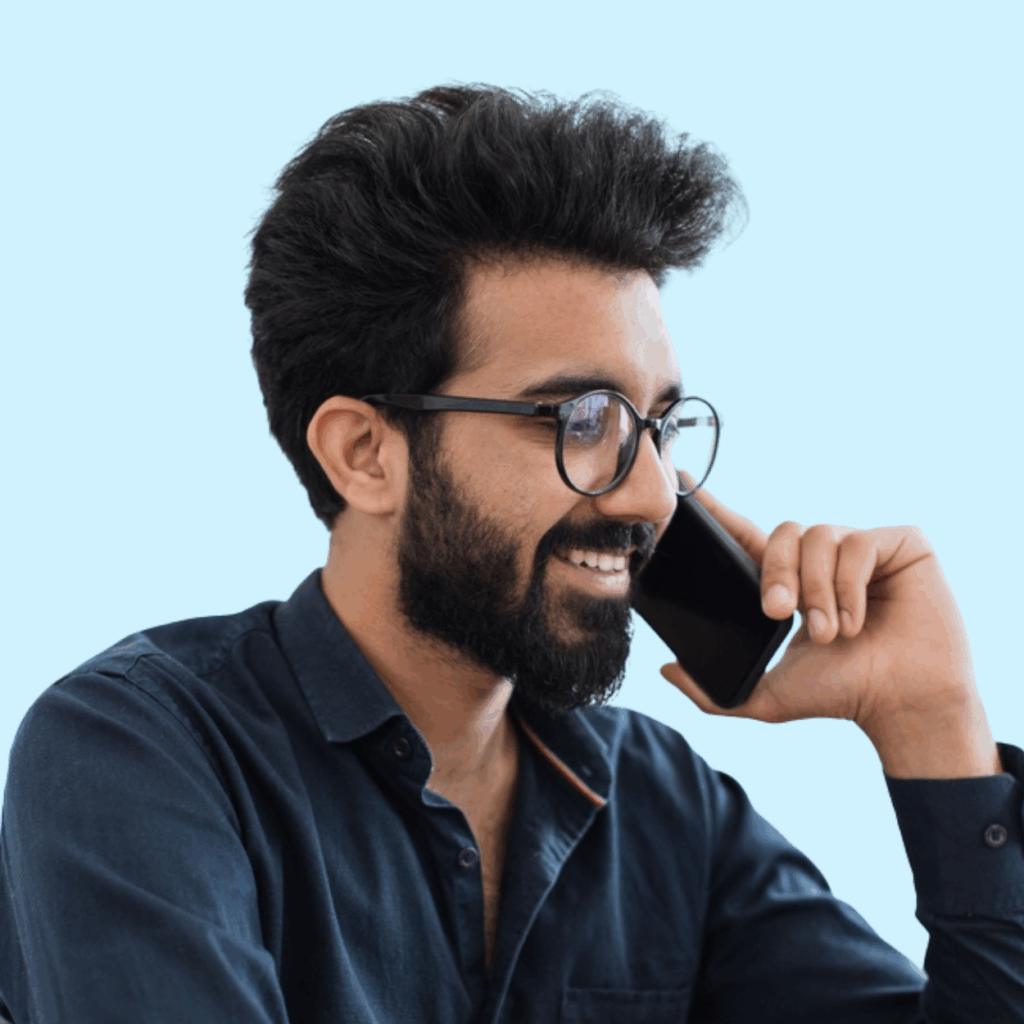 A man with dark hair, a beard, and glasses smiles while talking on a smartphone. He is wearing a dark shirt and is set against a light blue background.