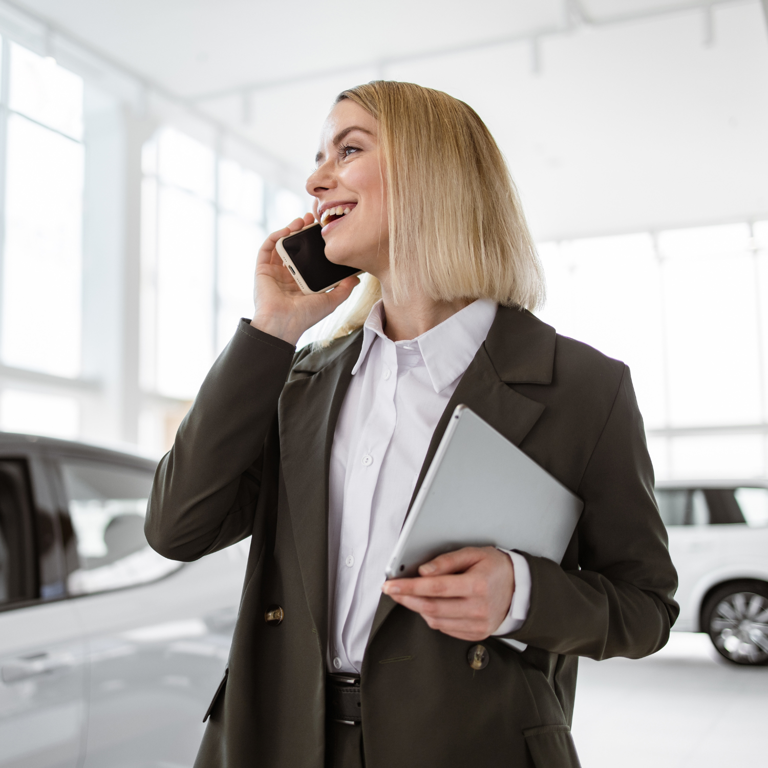 Smiling saleswoman smartphone while having business call at car showroom. Car dealer holding tablet pc in automobile showroom. Professional confident sales person working in modern car dealership.