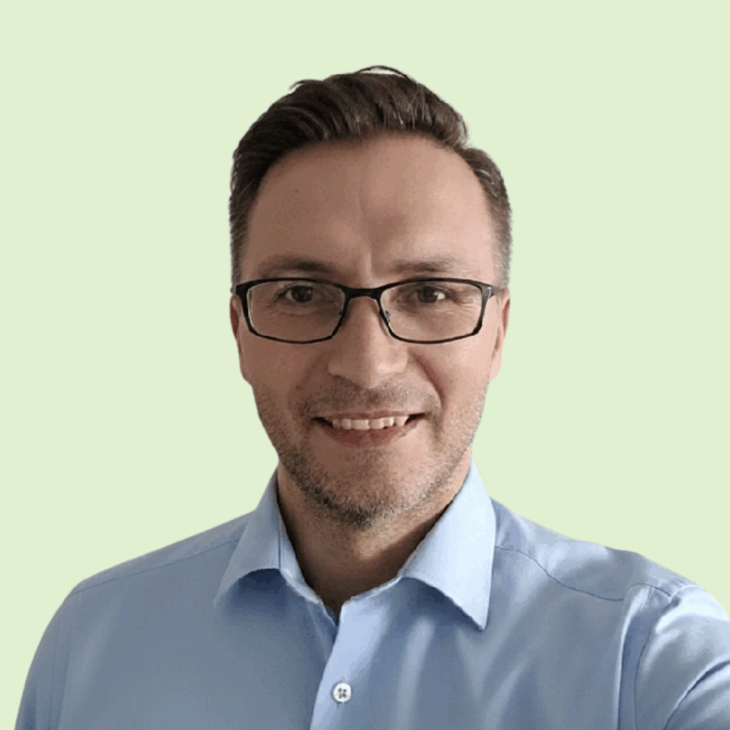A man with short brown hair and glasses is smiling, wearing a light blue button-up shirt, standing in front of a plain light green background.