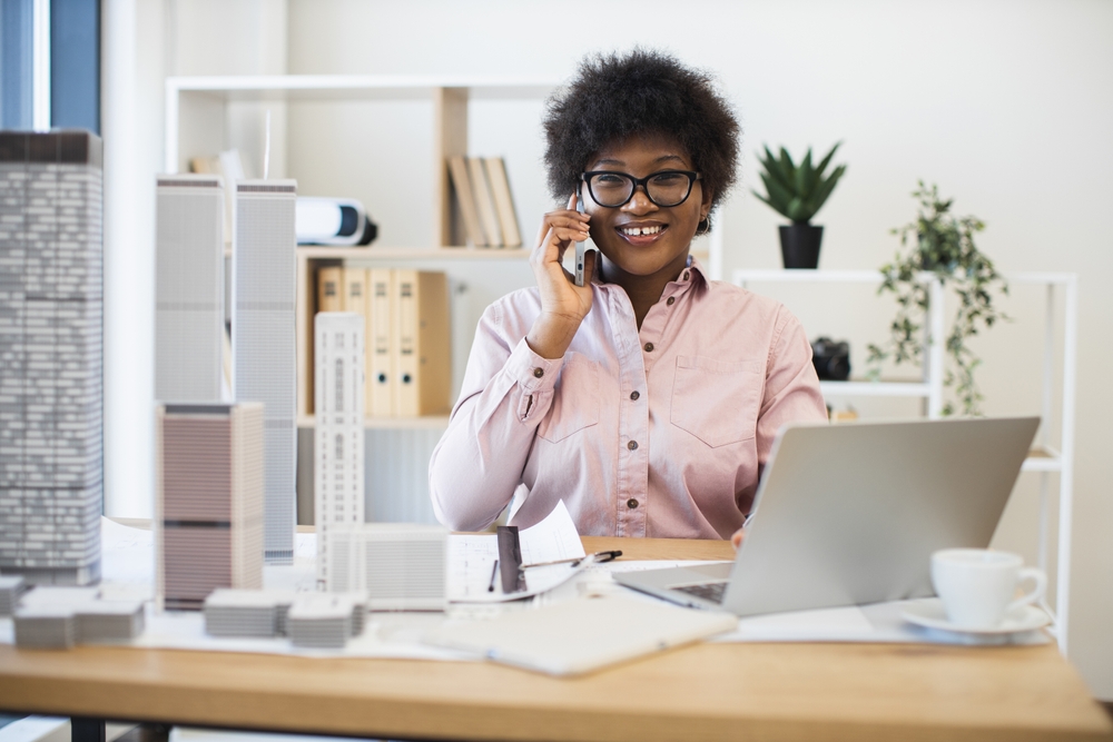 A woman wearing glasses and a pink shirt smiles while talking on the phone at her desk, surrounded by architectural models, a laptop, and paperwork in a modern office setting.