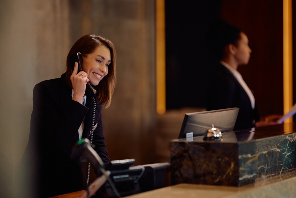 A smiling woman in professional attire talks on the phone at a hotel reception desk, with a computer monitor and service bell in front of her; another person stands blurred in the background.